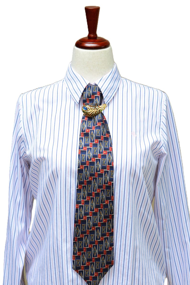 May include: A long-sleeved white button-down shirt with thin blue and red vertical stripes, paired with a dark blue necktie featuring a repeating geometric pattern of diamonds and red rectangles, secured by a gold-toned feather-shaped tie clip.