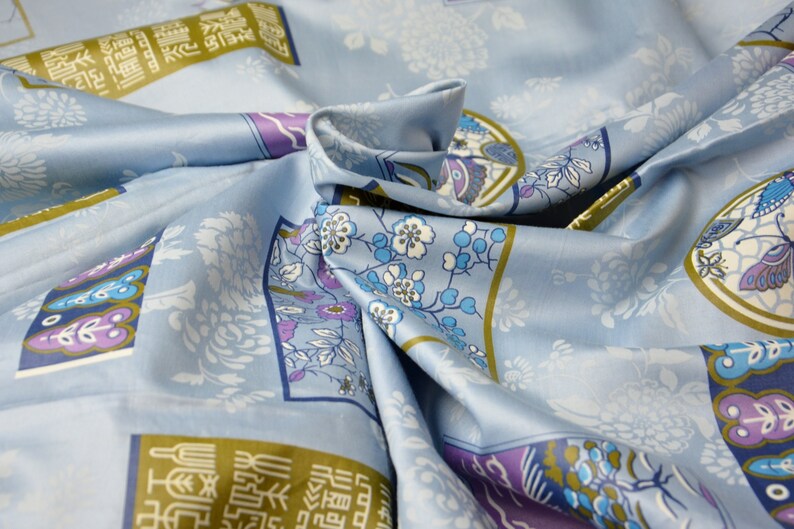 May include: Blue fabric with a repeating pattern of flowers, butterflies, and Chinese characters. The fabric is folded and draped, showing the pattern in detail.