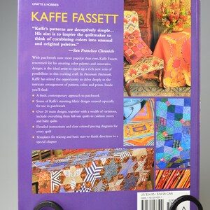 May include: A purple book titled "KAFFE FASSETT" on crafts and hobbies, displayed in a decorative black metal stand. The book cover features text about patchwork and colourful designs, with images of quilts.