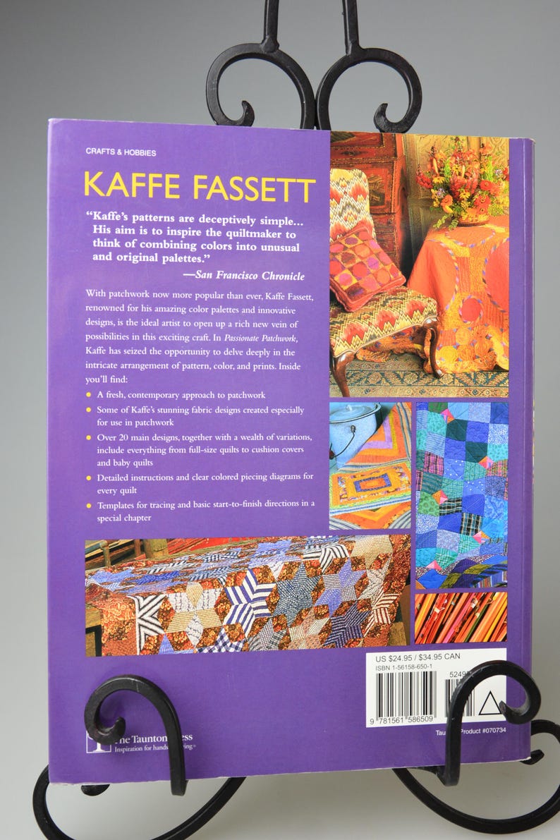 May include: A purple book titled "KAFFE FASSETT" on crafts and hobbies, displayed in a decorative black metal stand. The book cover features text about patchwork and colourful designs, with images of quilts.