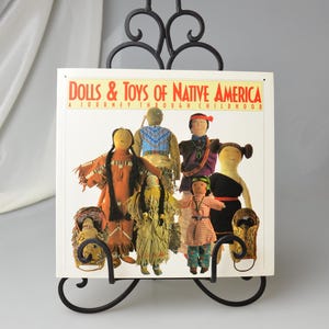 Pode incluir: Um livro intitulado "Dolls & Toys of Native America" exibido em um suporte de metal preto. A capa do livro apresenta ilustrações de várias bonecas nativas americanas com roupas tradicionais. O suporte tem volutas decorativas.