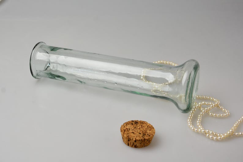 May include: A clear glass vase with a wide opening and a cork stopper. A strand of pearls is draped over the vase. The vase is lying on its side on a white surface. The vase is approximately 10 inches long.