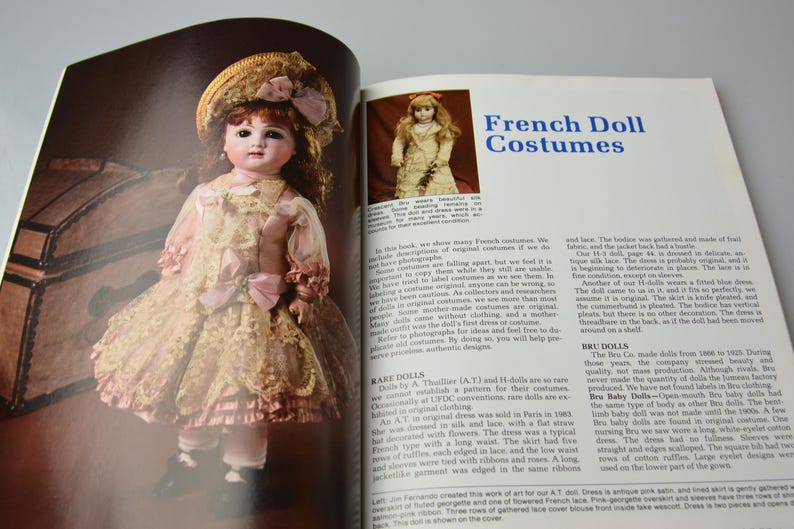 Pode incluir: Uma p&aacute;gina de livro vintage com uma foto de uma boneca de porcelana vestindo um vestido de renda branca e um chap&eacute;u de palha. O texto na p&aacute;gina diz "Trajes de Bonecas Francesas".