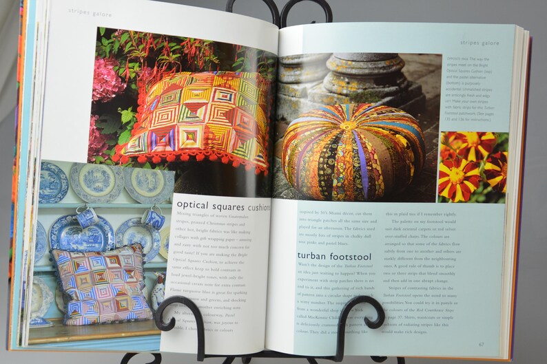 May include: An open book displaying images of a decorative pillow and a turban footstool. The pillow features a geometric design in red, orange, and yellow. The footstool is round with a striped pattern.