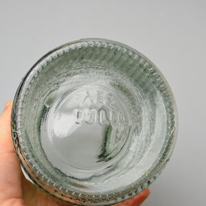 May include: A clear glass bottle base featuring a circular design with a textured, ribbed outer edge. The bottom of the glass displays embossed lettering that reads "AB" followed by "1010" in a stacked arrangement, centered within the circular glass base.