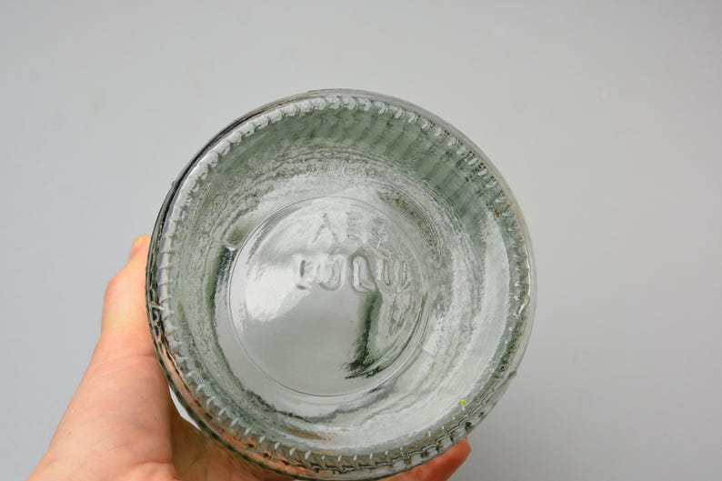 May include: A clear glass bottle base featuring a circular design with a textured, ribbed outer edge. The bottom of the glass displays embossed lettering that reads "AB" followed by "1010" in a stacked arrangement, centered within the circular glass base.