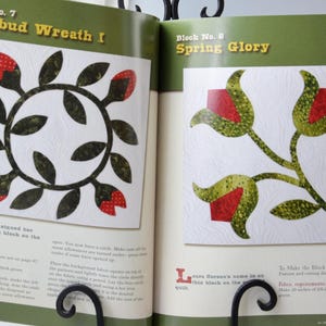 Może przedstawiać: Dwie strony książki o patchworku przedstawiające dwa r&oacute;żne wzory blok&oacute;w patchworkowych. Pierwszy blok nazywa się "Rosebud Wreath I" i zawiera wieniec z zielonych liści z czerwonymi pąkami r&oacute;ż. Drugi blok nazywa się "Spring Glory" i zawiera zieloną łodygę z trzema czerwonymi pąkami r&oacute;ż.