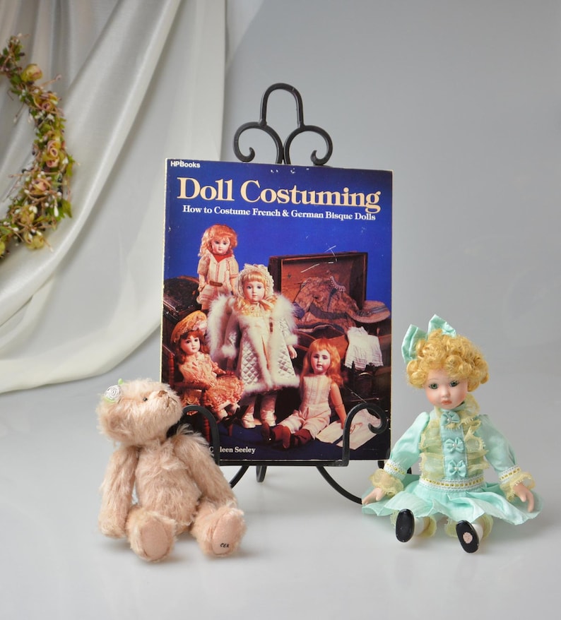 Pode incluir: Um livro intitulado "Doll Costuming: How to Costume French & German Bisque Dolls" de Jean Seeley &eacute; exibido em um suporte de metal preto. Um urso de pel&uacute;cia e uma boneca de porcelana est&atilde;o sentados na frente do livro.