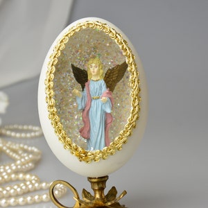 Puede incluir: Un huevo blanco con un borde dorado, que contiene una figurita de un &aacute;ngel con alas, dentro de una c&uacute;pula transparente llena de purpurina. El huevo est&aacute; sobre un soporte dorado con tres patas.