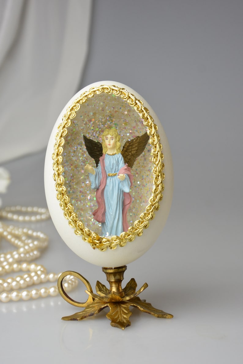 Puede incluir: Un huevo blanco con un borde dorado, que contiene una figurita de un &aacute;ngel con alas, dentro de una c&uacute;pula transparente llena de purpurina. El huevo est&aacute; sobre un soporte dorado con tres patas.
