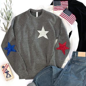 以下が含まれることがあります： グレーのトレーナーに、前面に白い星、袖に赤と青の星がデザインされています。トレーナーは青いジーンズと合わせています。