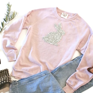 Könnte beinhalten: Ein rosa Sweatshirt mit einem silbernen Pailletten-Hasen-Design. Das Sweatshirt wird mit einer blauen Jeans und einer Sonnenbrille kombiniert.