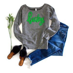 May include: Gray sweatshirt with a green glitter "Lucky" graphic. The sweatshirt is paired with blue jeans and black fuzzy slippers.