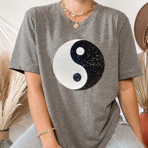 Pode incluir: Uma camiseta cinza com um símbolo yin yang preto e branco feito de lantejoulas. A camiseta é oversized e tem um caimento relaxado.