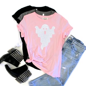 Op de afbeelding: Een roze T-shirt met een wit glitter-spookontwerp. Het shirt wordt gecombineerd met een blauwe jeans en zwarte studded laarzen.