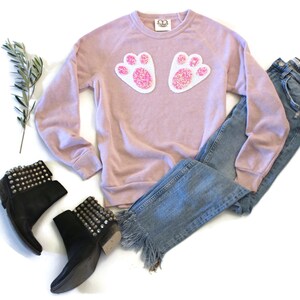 Könnte beinhalten: Ein staubiges rosafarbenes Sweatshirt mit pinkfarbenen und weißen Pailletten-Hasenpfoten auf der Vorderseite. Das Sweatshirt wird mit blauen Jeans und schwarzen, mit Nieten besetzten Stiefeletten kombiniert.