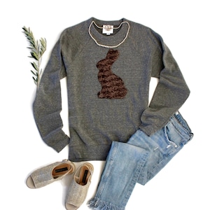 Könnte beinhalten: Ein graues Sweatshirt mit einer braunen Hasensilhouette auf der Vorderseite. Das Sweatshirt wird mit einer blauen Jeans und beigen Sandalen kombiniert.