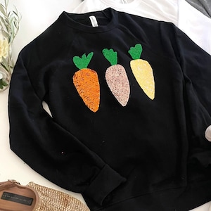 Könnte beinhalten: Schwarzer Pullover mit drei gestickten Karotten in Orange, Gelb und Rosa. Die Karotten haben grüne Blätter.
