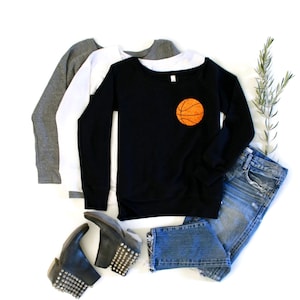 May include: A black long-sleeved sweatshirt with a basketball graphic on the front. The sweatshirt is layered over a white long-sleeved shirt and a grey long-sleeved shirt. The sweatshirt is paired with blue jeans and black studded ankle boots.