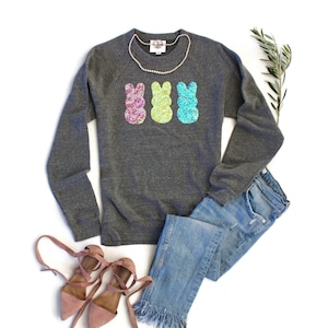 Könnte beinhalten: Ein graues Langarmshirt mit einem Aufdruck von drei pastellfarbenen Osterhasen. Das Shirt wird mit einer blauen Jeans und rosa Sandalen kombiniert.