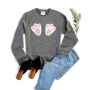 Könnte beinhalten: Ein grauer Pullover mit einem weißen und rosafarbenen Hasenpfoten-Design. Der Pullover wird mit einer blauen Jeans und schwarzen Schuhen gezeigt.