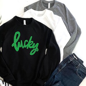 Może przedstawiać: Czarna bluza z zielonym brokatowym napisem "lucky". Bluza jest połączona z niebieskimi dżinsami i białymi butami sportowymi.
