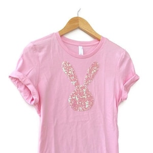 Könnte beinhalten: Ein pinkfarbenes T-Shirt mit einem Pailletten-Hasendesign auf der Vorderseite.
