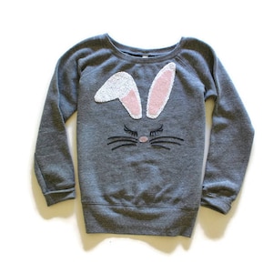 May include: Gray sweatshirt with a bunny face design. The bunny ears are white with pink inner ears, and the face features black whiskers and eyelashes. A cute and festive top for Easter or everyday wear.