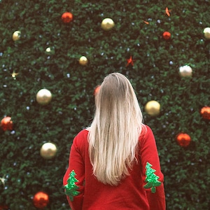 Puede incluir: Una persona con un suéter rojo con árboles de Navidad verdes en las mangas se encuentra frente a un gran árbol de Navidad decorado con adornos rojos y dorados.