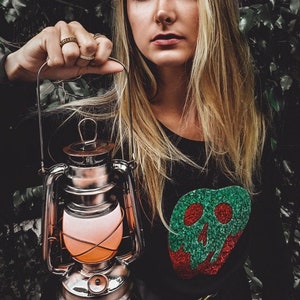 May include: A woman with long blonde hair holds a silver lantern with a red and orange glow. She is wearing a black sweater with a green and red glitter apple design.