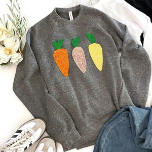 Könnte beinhalten: Ein grauer Pullover mit drei gestickten Karotten in Orange, Rosa und Gelb. Die Karotten haben grüne Blätter.