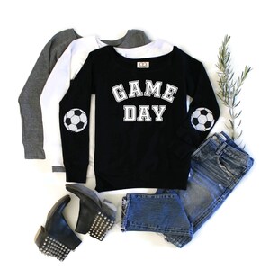 以下が含まれることがあります： 「GAME DAY」の白い文字と肘にサッカーボールのパッチが付いた黒い長袖スウェットシャツ。画像には、グレーと白のスウェットシャツ、ブルージーンズ、黒いスタッズ付きブーツも写っています。