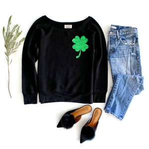 May include: Black sweatshirt with a green four-leaf clover sequin design. The sweatshirt is paired with blue jeans and black fuzzy slippers.