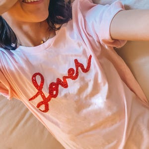 Puede incluir: Una camiseta rosa con la palabra "lover" escrita en purpurina roja.