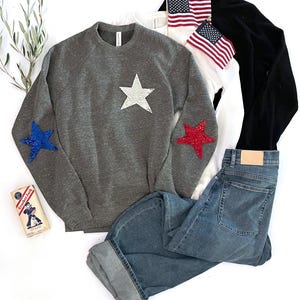 Puede incluir: Una sudadera gris con una estrella blanca en la parte delantera y estrellas rojas y azules en las mangas. La sudadera se combina con unos vaqueros azules.