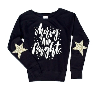May include: Black sweatshirt with long sleeves and gold glitter star patches on the elbows. The front of the sweatshirt has the text "Merry and Bright" in white.