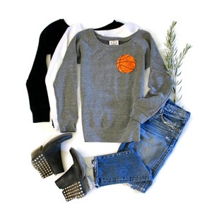 May include: A grey sweatshirt with a basketball graphic on the front. The sweatshirt is paired with blue jeans and black studded ankle boots.