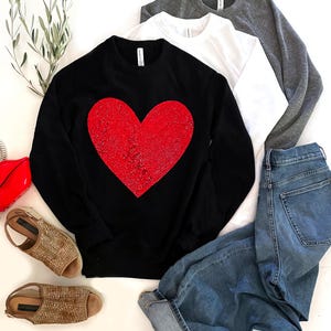 May include: A black sweatshirt with a large red glitter heart on the front. The sweatshirt is shown with a pair of jeans and sandals.