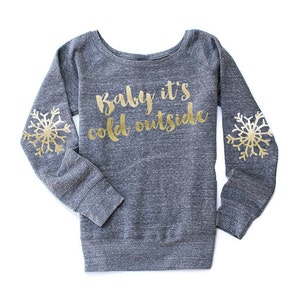 May include: A grey sweatshirt featuring the gold text "Baby it's cold outside." Gold snowflake designs adorn each sleeve. The sweatshirt has a scoop neck and long sleeves. The fabric has a heathered appearance.