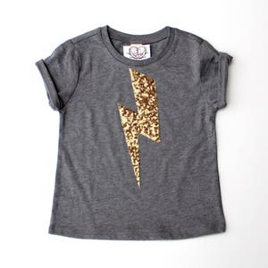 Puede incluir: Una camiseta gris de manga corta con un diseño de rayo dorado de lentejuelas.