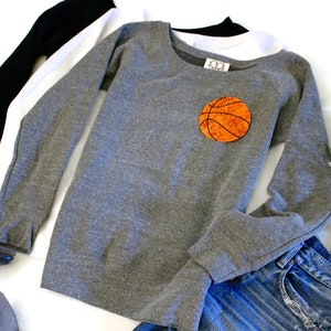 May include: Grey sweatshirt with long sleeves and a round orange basketball patch on the front. The sweatshirt is laid out on a white surface with a pair of jeans and a pair of boots.