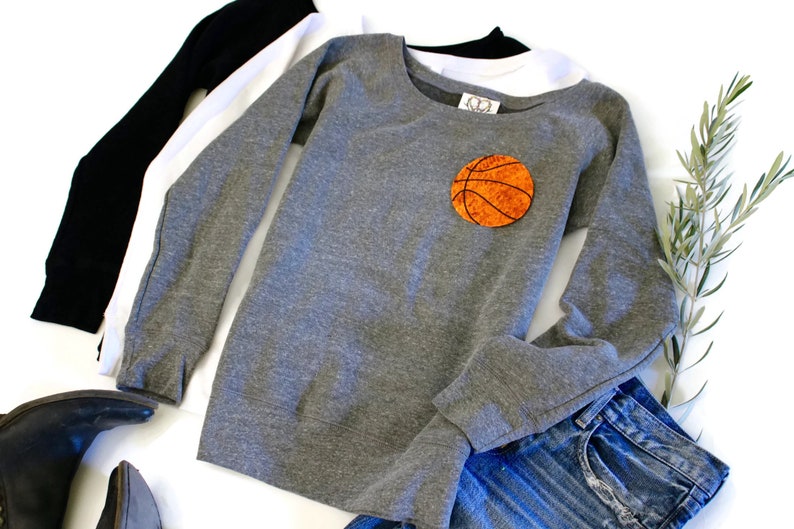 May include: Grey sweatshirt with long sleeves and a round orange basketball patch on the front. The sweatshirt is laid out on a white surface with a pair of jeans and a pair of boots.