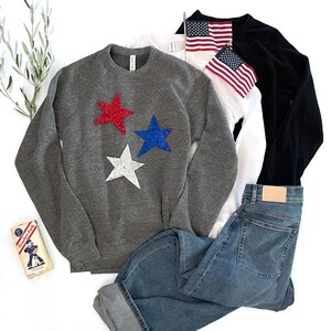 May include: A gray sweatshirt with three glitter stars in red, white, and blue. The stars are arranged in a cluster on the front of the sweatshirt. A pair of blue jeans is also pictured.
