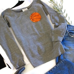 May include: A grey sweatshirt with a basketball graphic on the front. The sweatshirt is laid out on a white surface with a pair of blue jeans and a white and black long-sleeved shirt.