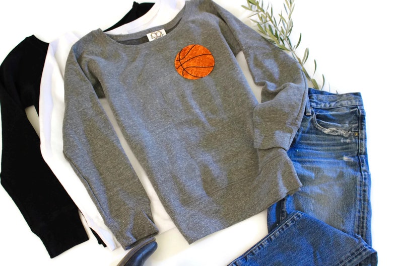 May include: A grey sweatshirt with a basketball graphic on the front. The sweatshirt is laid out on a white surface with a pair of blue jeans and a white and black long-sleeved shirt.