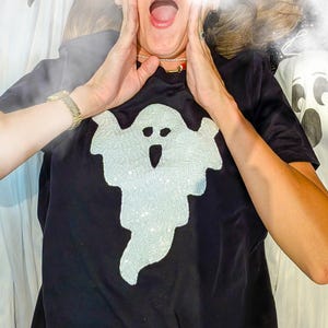 Op de afbeelding: Een zwart t-shirt met korte mouwen en een grote, centrale afbeelding van een geest. Het ontwerp van de geest is gevuld met glinsterende, lichtgekleurde pailletten, wat zorgt voor een textuur op de effen zwarte stof van het shirt.