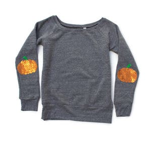 Peut inclure: Un sweat-shirt gris avec des manches longues et un col rond. Les coudes présentent des patchs de citrouilles orange à paillettes avec des tiges vertes. Le sweat-shirt est posé à plat sur une surface blanche, mettant en valeur un design sur le thème de l'automne.