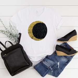 May include: White t-shirt with a gold and black crescent moon design. The image also includes a black backpack, blue jeans, and black wedge heels. The items are arranged on a white wooden surface.