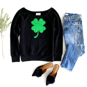 May include: Black sweatshirt with a green four-leaf clover graphic. The sweatshirt is paired with blue jeans and black fuzzy slippers.
