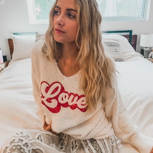May include: A white fuzzy sweater with a red "Love" graphic. The sweater is worn by a person sitting on a bed with a white bedspread. The person is wearing a white skirt with a lace design.